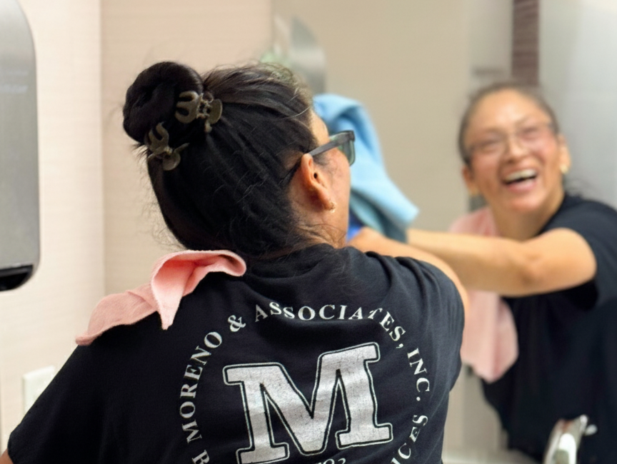 Moreno janitor smiling while cleaning a mirror with a blue rag
